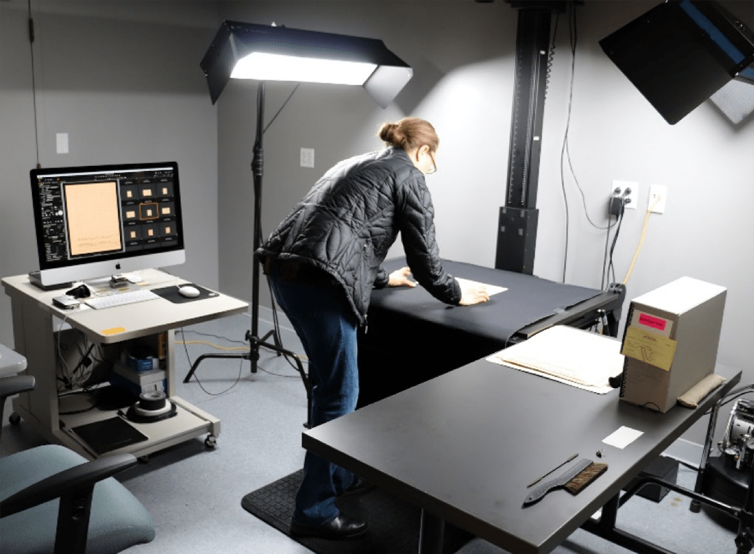 Cheryl Ostrowski, Digital Content Specialist, adjusts a page from the Anansi stories on a flat black surface to capture a digital image with the Phase One Camera (out of sight at the top of this photo, because of the raised ceiling). The computer to the left shows recently digitized images.