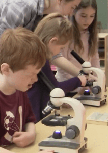 Three children look through lenses of a microscope while a teacher assists them.